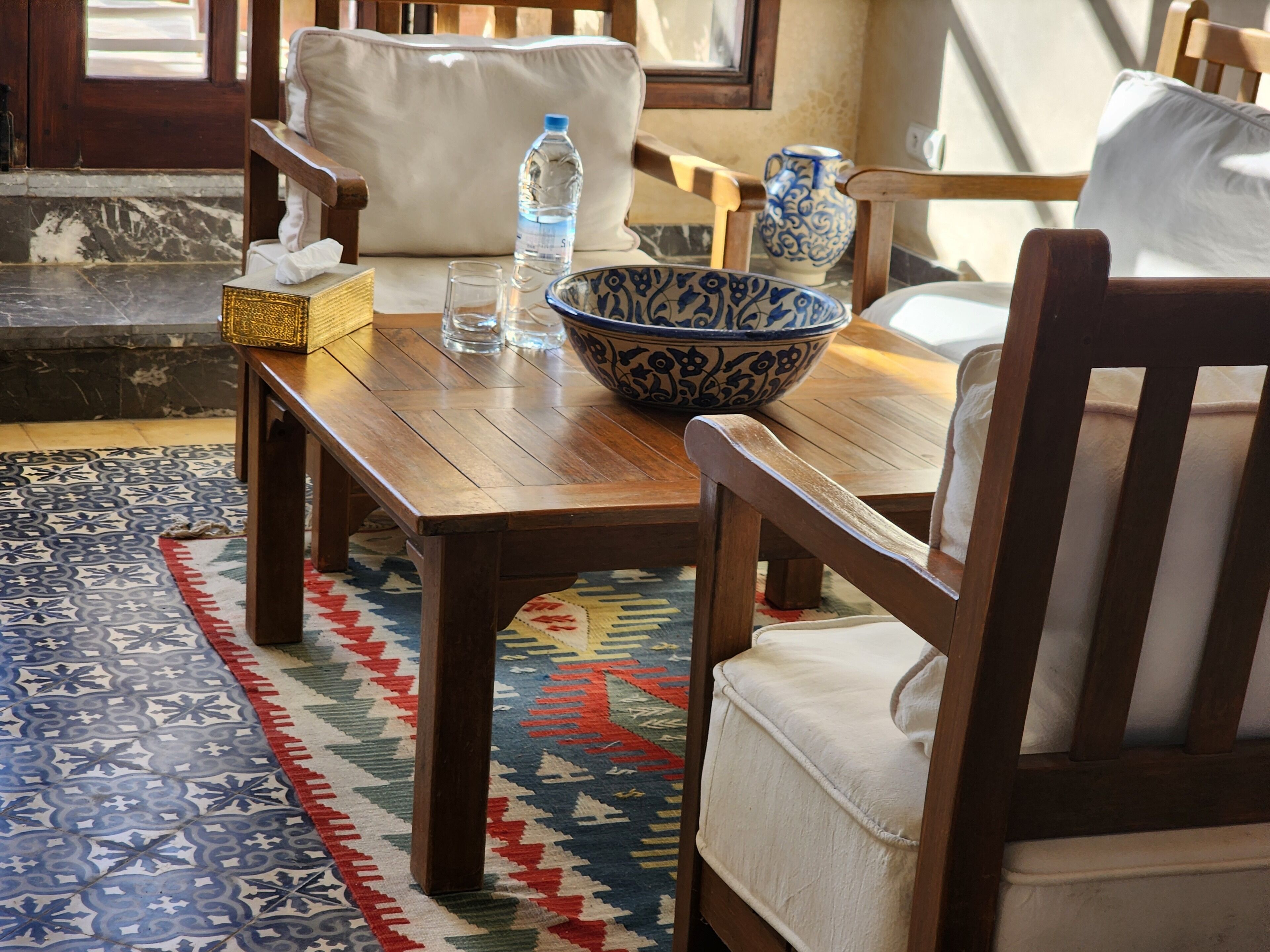 A wooden table with a bowl and glasses, a patterned rug, and a chair with a cushion.