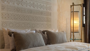 Una cama bien hecha con almohadas blancas y marrones, una pared decorativa con diseños intrincados y una lámpara alta y estilizada fijada a la pared.