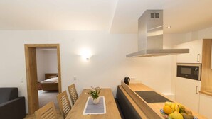 Un espace de vie moderne et compact, comprenant une table à manger en bois, un banc, un coin cuisine et une petite chambre à coucher visible à travers une porte ouverte.