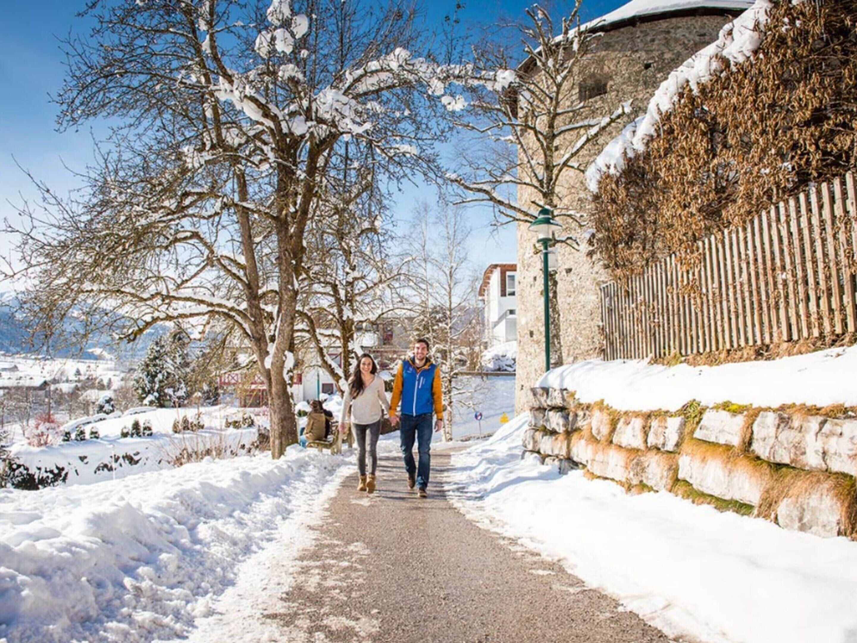 A snow-covered pathway with a stone wall on one side and bare trees on the other.