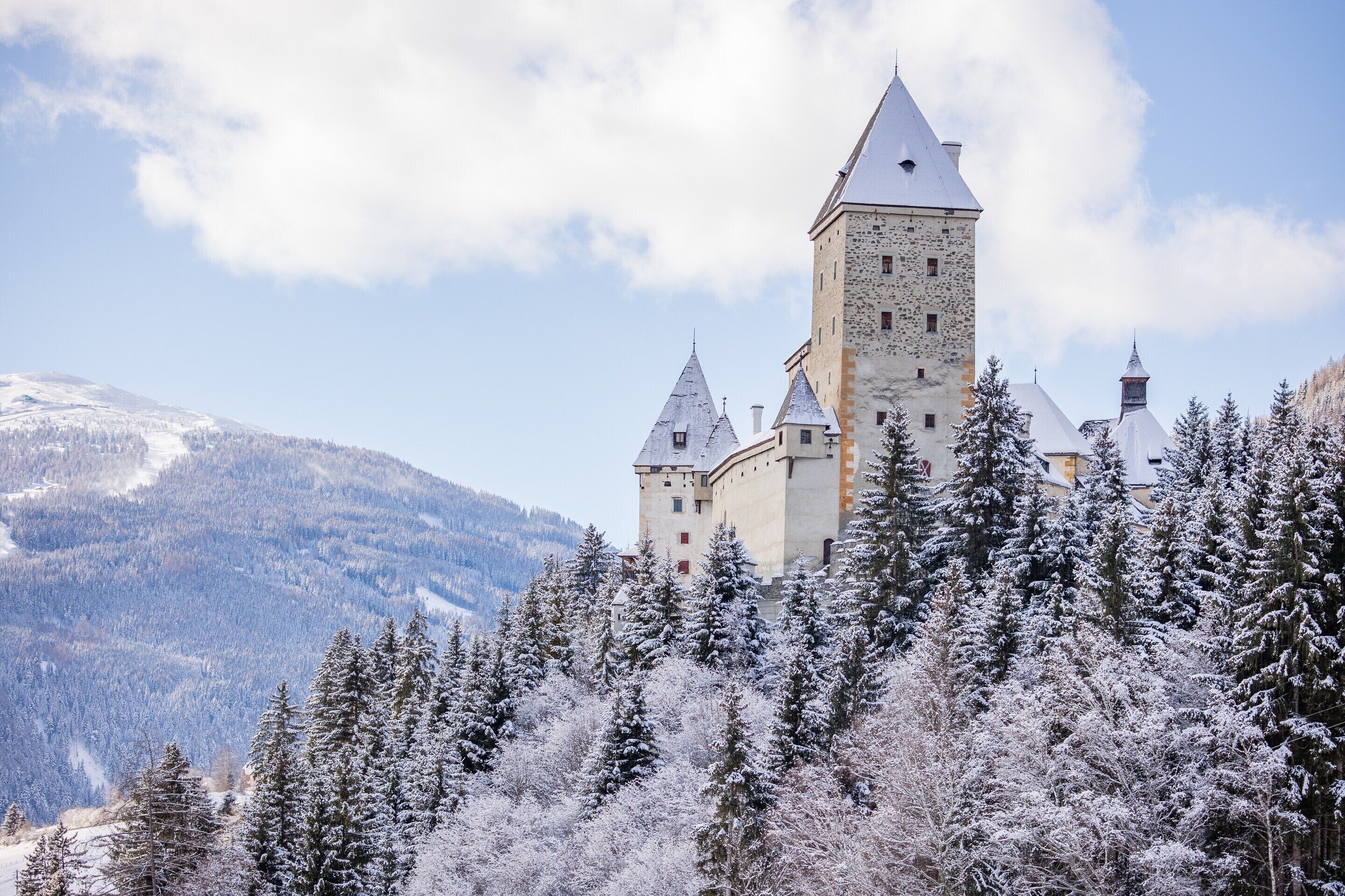 Un château doté de nombreuses tours, avec une tour centrale particulièrement imposante, entouré d’arbres et de montagnes couverts de neige.