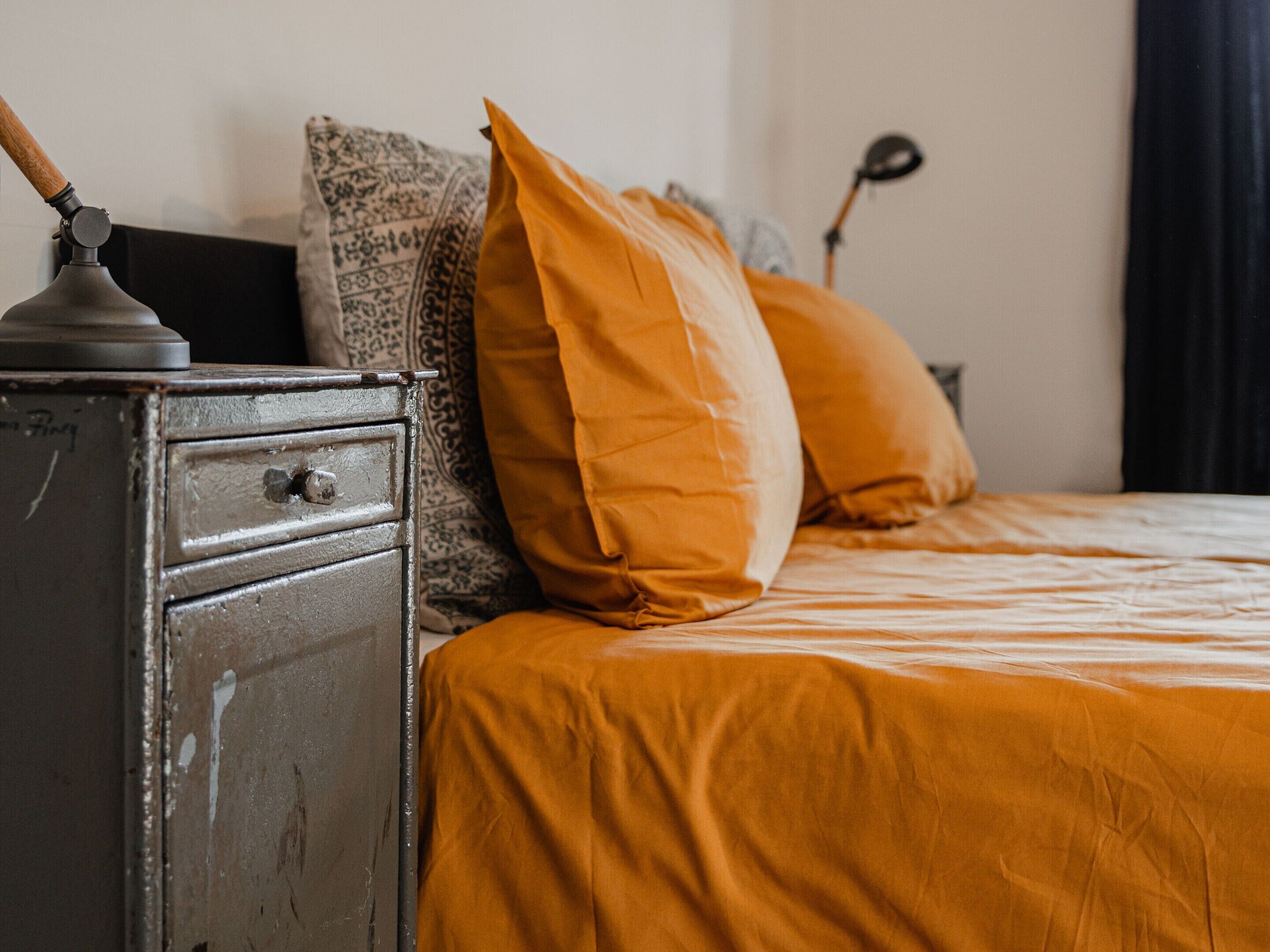 Una cama con ropa de cama naranja, un cabecero de madera y un aparador metálico con cajones.