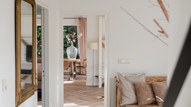 A room with a checkered floor, a wooden sofa with cushions, a mirror, and a view into another room with a table and chairs.