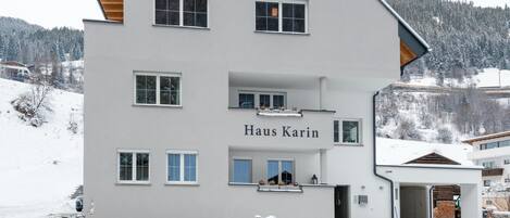 A multi-story building named Haus Karin, featuring a stone wall in front, multiple windows, and a snowy landscape in the background.