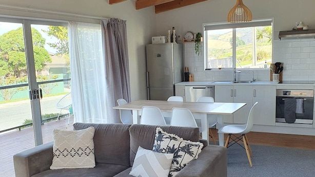 Un salon moderne avec un canapé gris, des chaises blanches et une table basse en bois. On aperçoit, au fond, une cuisine avec des meubles blancs et un luminaire suspendu.