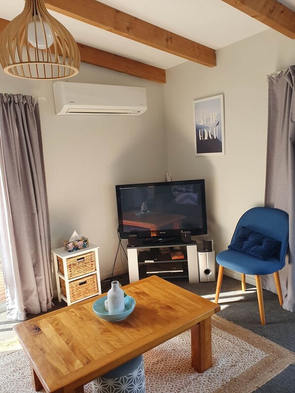 En stue med et træsofabord, en blå lænestol, et fladskærms-tv og en vægmonteret aircondition.