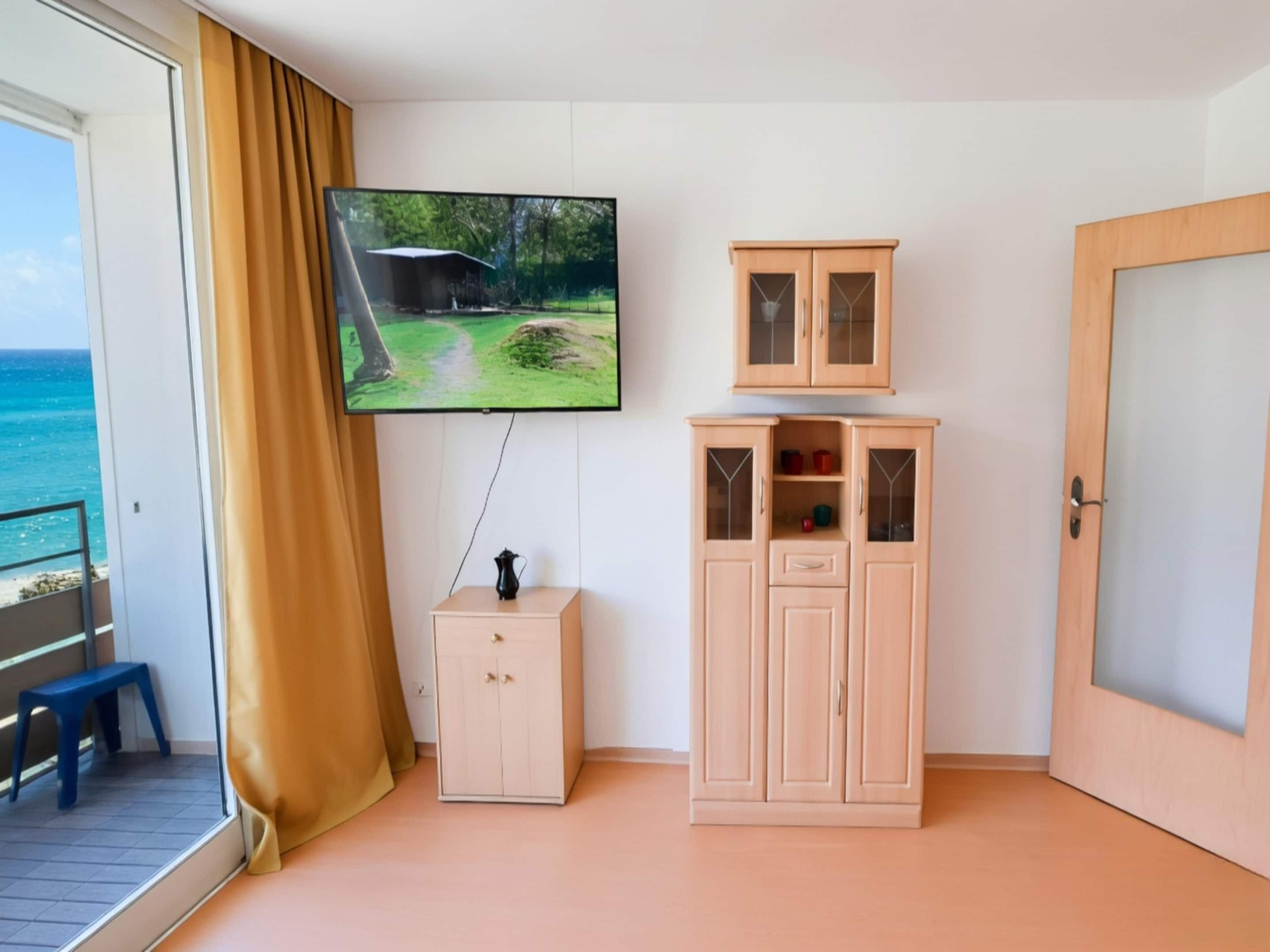 Huoneessa on seinälle asennettu televisio, puinen kaappi, pieni pöytä, jossa on kannullinen juomaa, ja liukuoven kautta avautuu näkymä merelle.