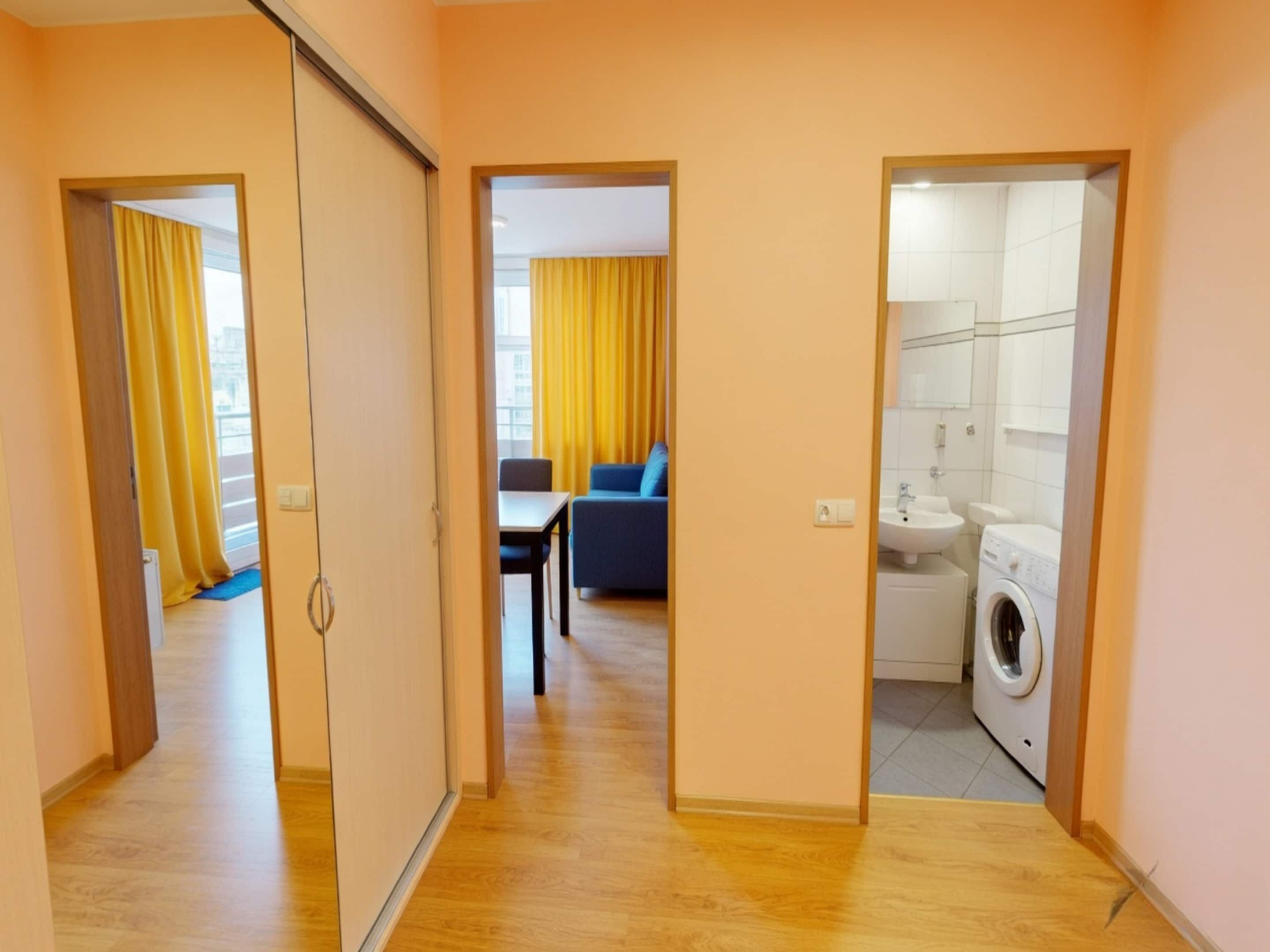 Un couloir mène à un espace de vie comprenant un canapé et une table à manger, ainsi qu’à une pièce séparée où se trouve une machine à laver.