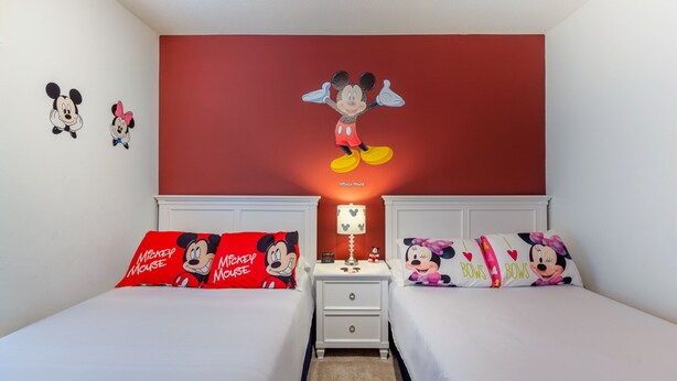 Una habitación de hotel con dos camas, cada una con ropa de cama temática de Mickey Mouse. Hay una mesita de noche entre las camas con una lámpara de Mickey Mouse.
