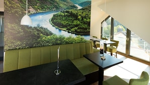 A modern dining area with a large scenic mural, green upholstered seating, and a view of a river and forest through large windows. - Modern Apartment in Mettlach With Infrared Sauna (Mettlach)