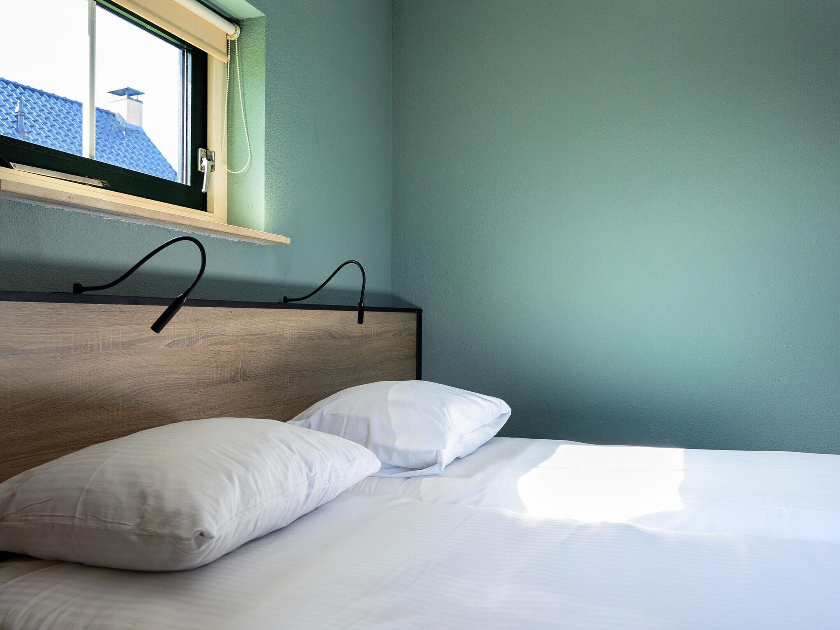 A bedroom with a wooden headboard, two white pillows, and a window with a view of rooftops.