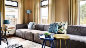 Een moderne woonkamer met een grijze bank, twee ronde tafels, een lamp en uitzicht op bomen door grote ramen.