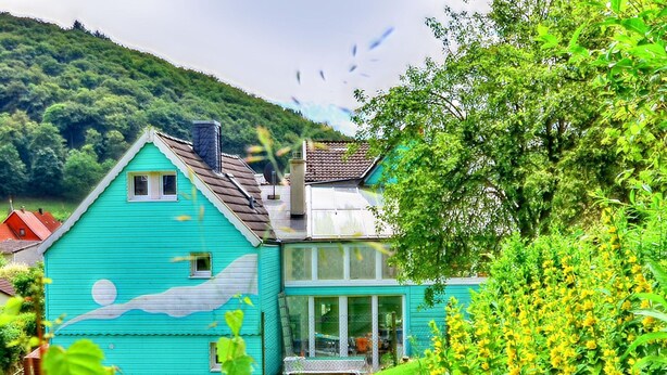 Una casa turquesa con un mural de piscina, rodeada de exuberante follaje y flores amarillas.