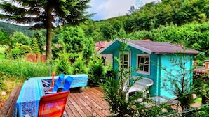 Una pequeña cabaña turquesa con terraza de madera, rodeada de exuberante follaje y árboles.
