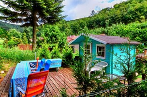 Una pequeña cabaña turquesa con terraza de madera, rodeada de exuberante follaje y árboles.