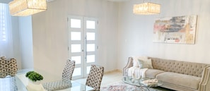 A modern living room with a glass coffee table, a beige sofa, patterned chairs, and a large abstract painting on the wall.