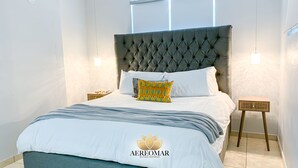 Un lit bien fait, doté d’une tête de lit capitonnée, de draps blancs et d’un coussin décoratif. Deux tables de chevet avec des lampes se trouvent de part et d’autre du lit, et la pièce présente une esthétique épurée et moderne.