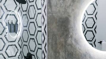 Un baño con inodoro blanco, lavamanos con dispensador de jabón dorado y ducha con patrón de baldosas hexagonales blanco y negro.