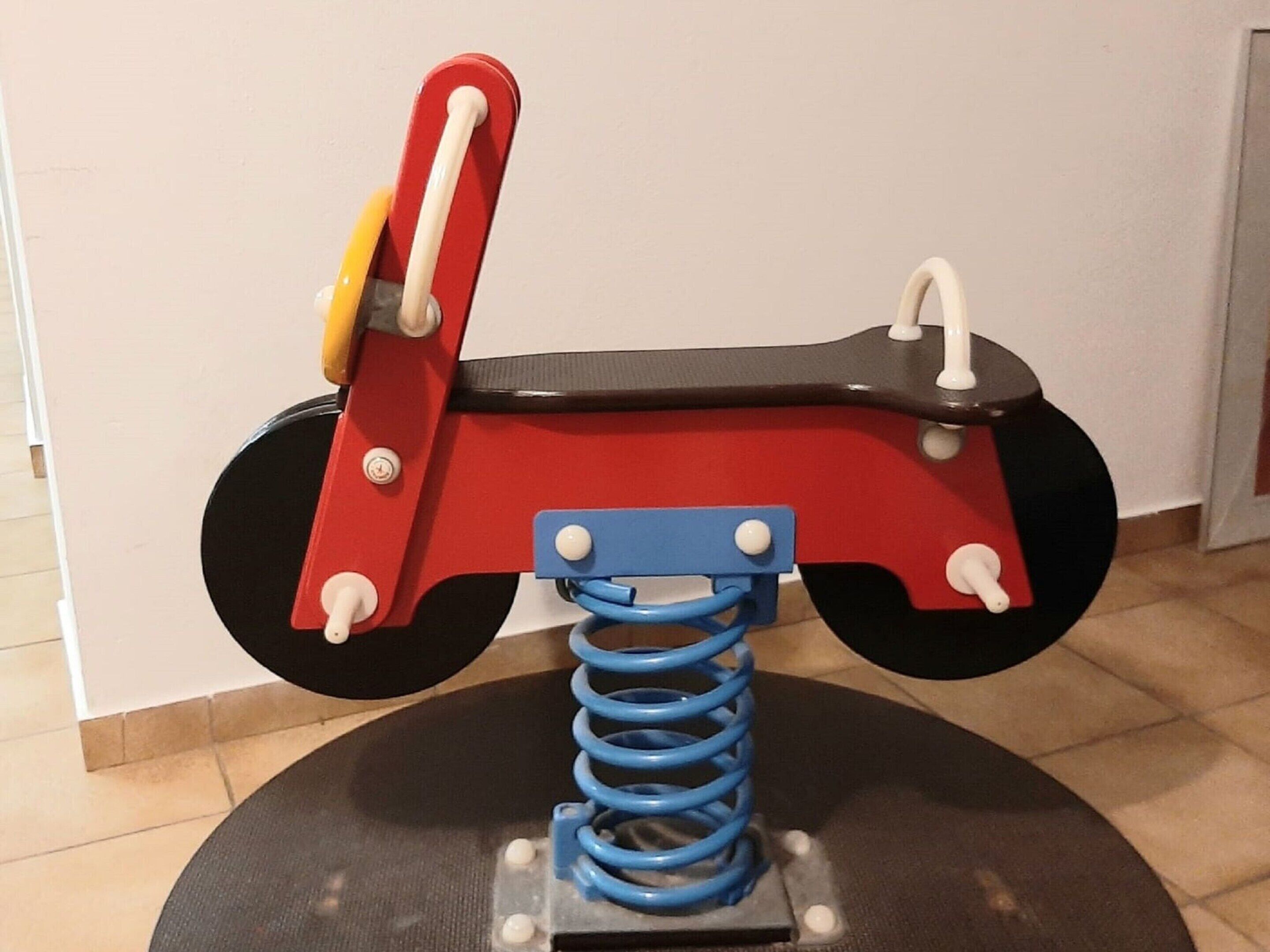 Ein rotes und schwarzes Spielzeugmotorrad mit blauer Federbasis, montiert auf einer schwarzen, runden Plattform.