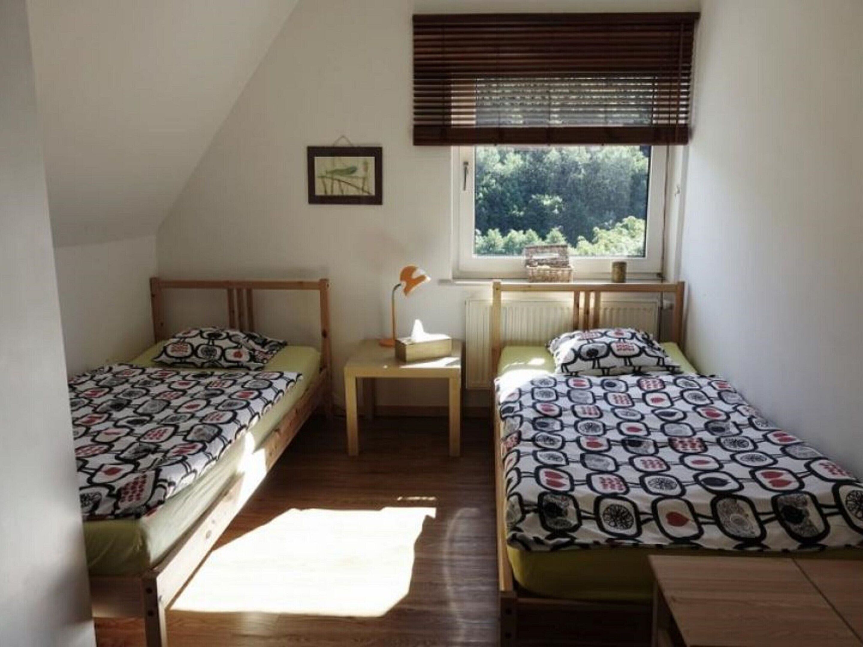 A small, well-lit room with two single beds, a nightstand, a window with blinds, and a framed picture on the wall.