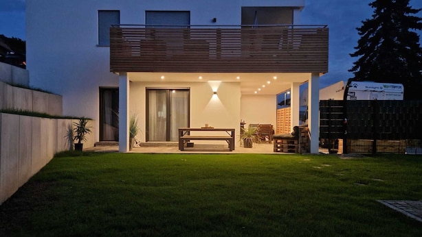 Een modern huis met twee verdiepingen, een balkon, een patio met een tafel en stoelen en een verzorgd gazon.