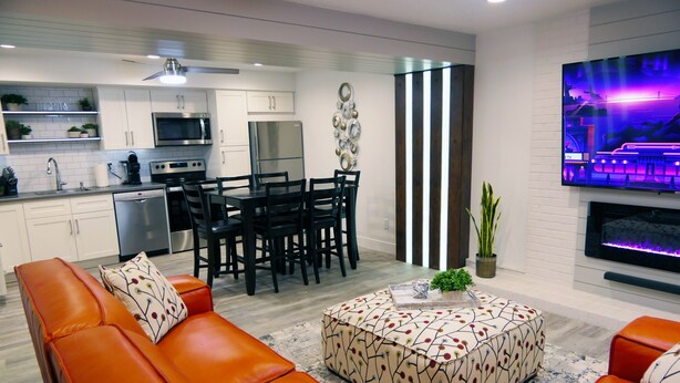 A modern kitchen with white cabinets, a microwave, and a refrigerator. There is a dining area with black chairs and a table. A large flat-screen TV is mounted on the wall. - Natures Best Modern - Amazing GETAWAY, 2- Person LED Shower, 75" TV, King Bed (Orem)