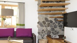 A modern living room with a stone wall, wooden staircase, a flat-screen TV, and a sofa with purple cushions.