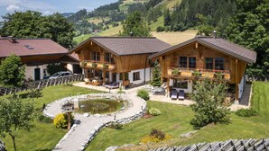 A wooden chalet with a pond, surrounded by a well-maintained garden and a view of rolling hills.
