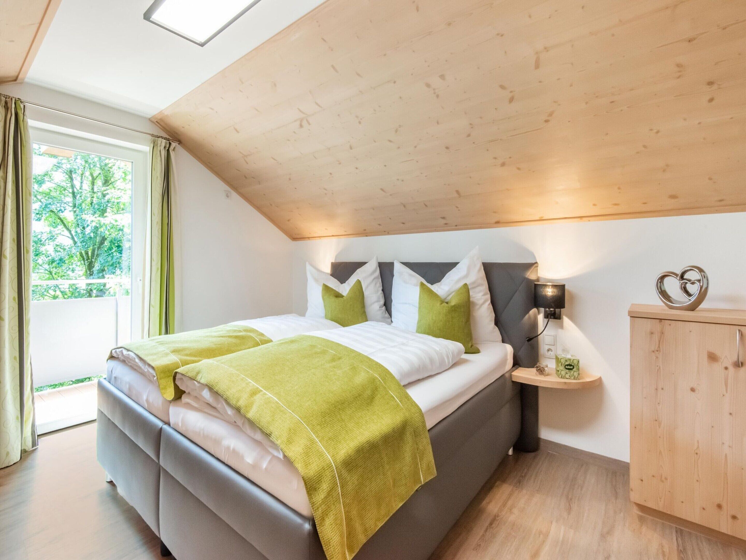 A modern bedroom with a large bed, a wooden ceiling, a nightstand with a lamp, and a view of greenery outside.