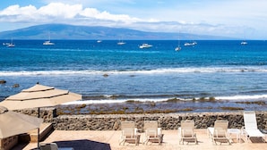 A beach with lounge chairs and umbrellas, a rocky shoreline, and sailboats on the water. - Lahaina Roads 206 - Complete remodel - Oceanfront - You can't beat the view and location! (Lahaina)