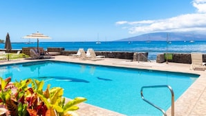 A pool with lounge chairs and umbrellas, a stone wall, and a view of the sea with boats. - Lahaina Roads 206 - Complete remodel - Oceanfront - You can't beat the view and location! (Lahaina)