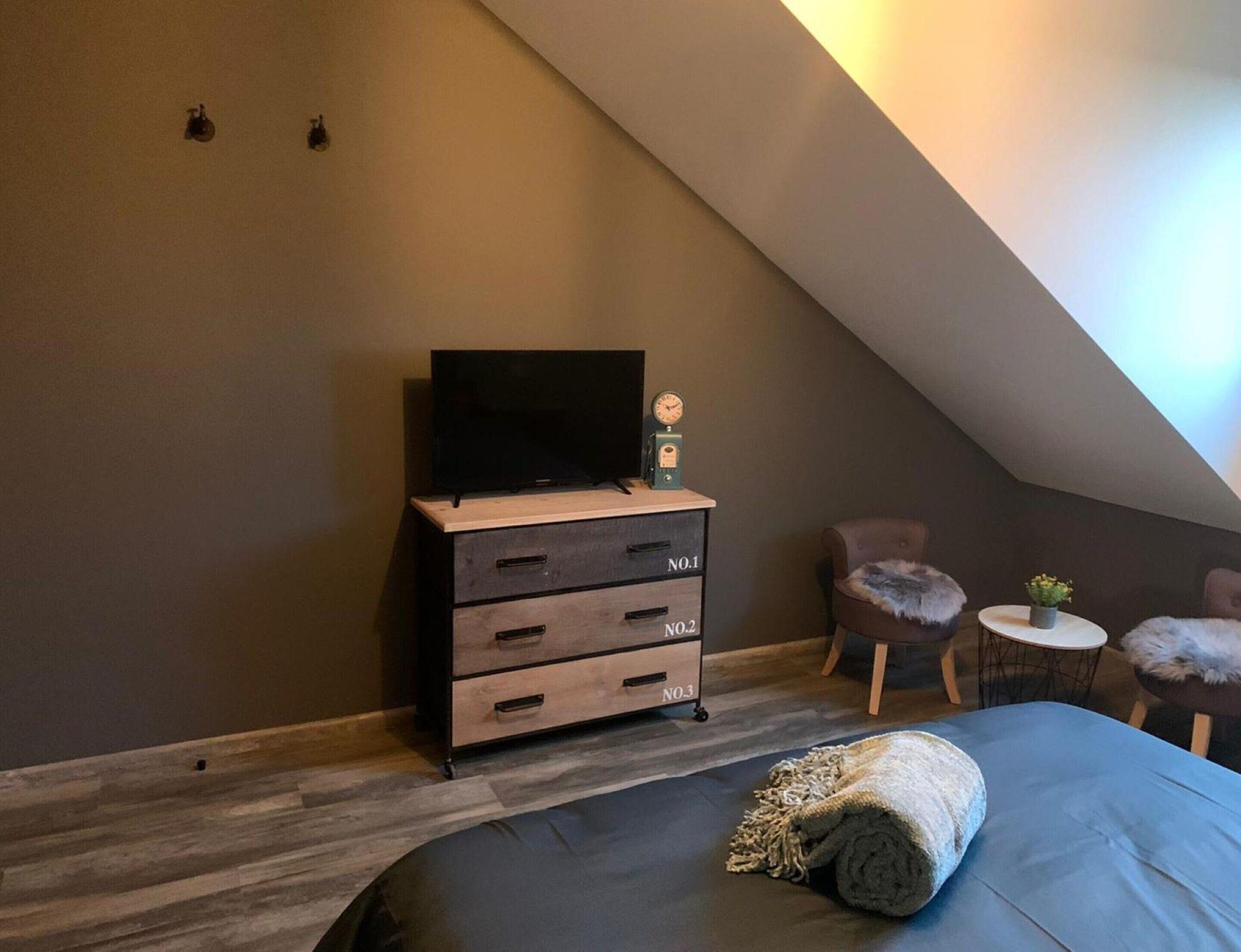 A bedroom with a sloped ceiling, a bed with a rolled-up blanket, a dresser with drawers, a television, and a small table with a plant.