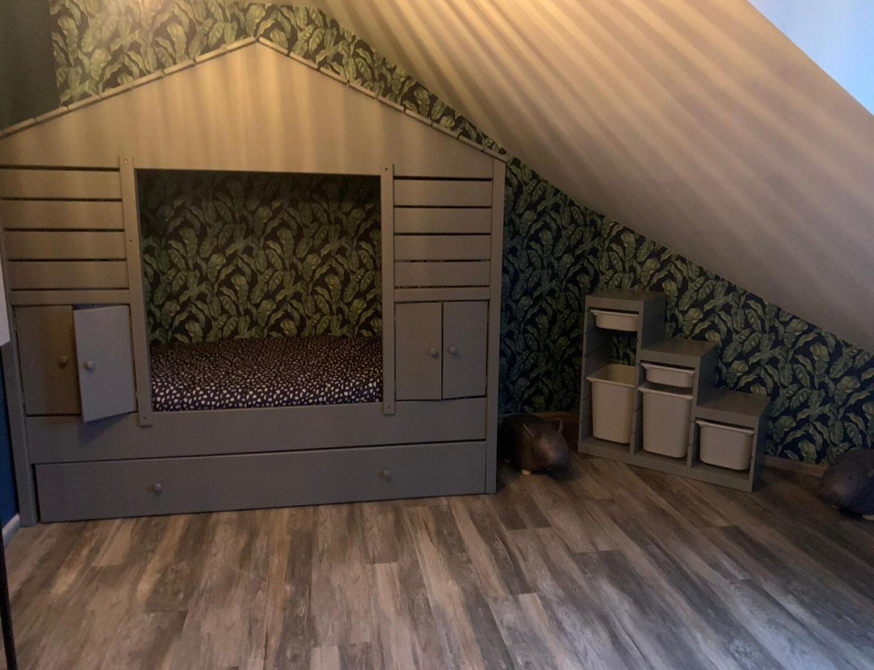 Een kamer met een schuine kap, een in het interieur geïntegreerd bed met opbergkasten, een tafeltje en behang met een patroon.