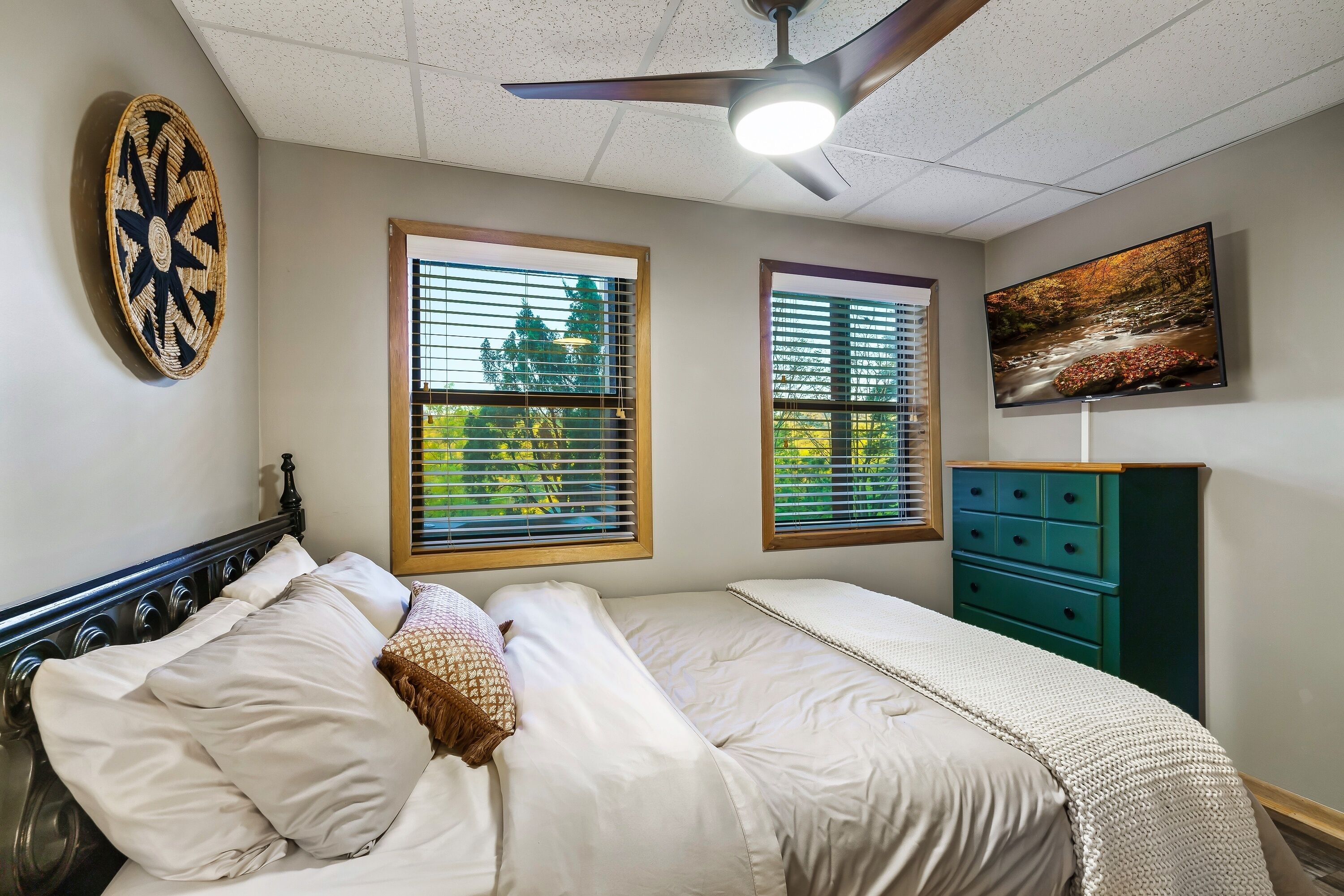 A bedroom with a bed, a ceiling fan, a wall-mounted TV, and two windows with blinds.