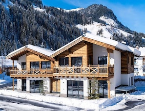 Un chalet de madera de dos pisos con balcón, frente a un paisaje montañoso nevado.