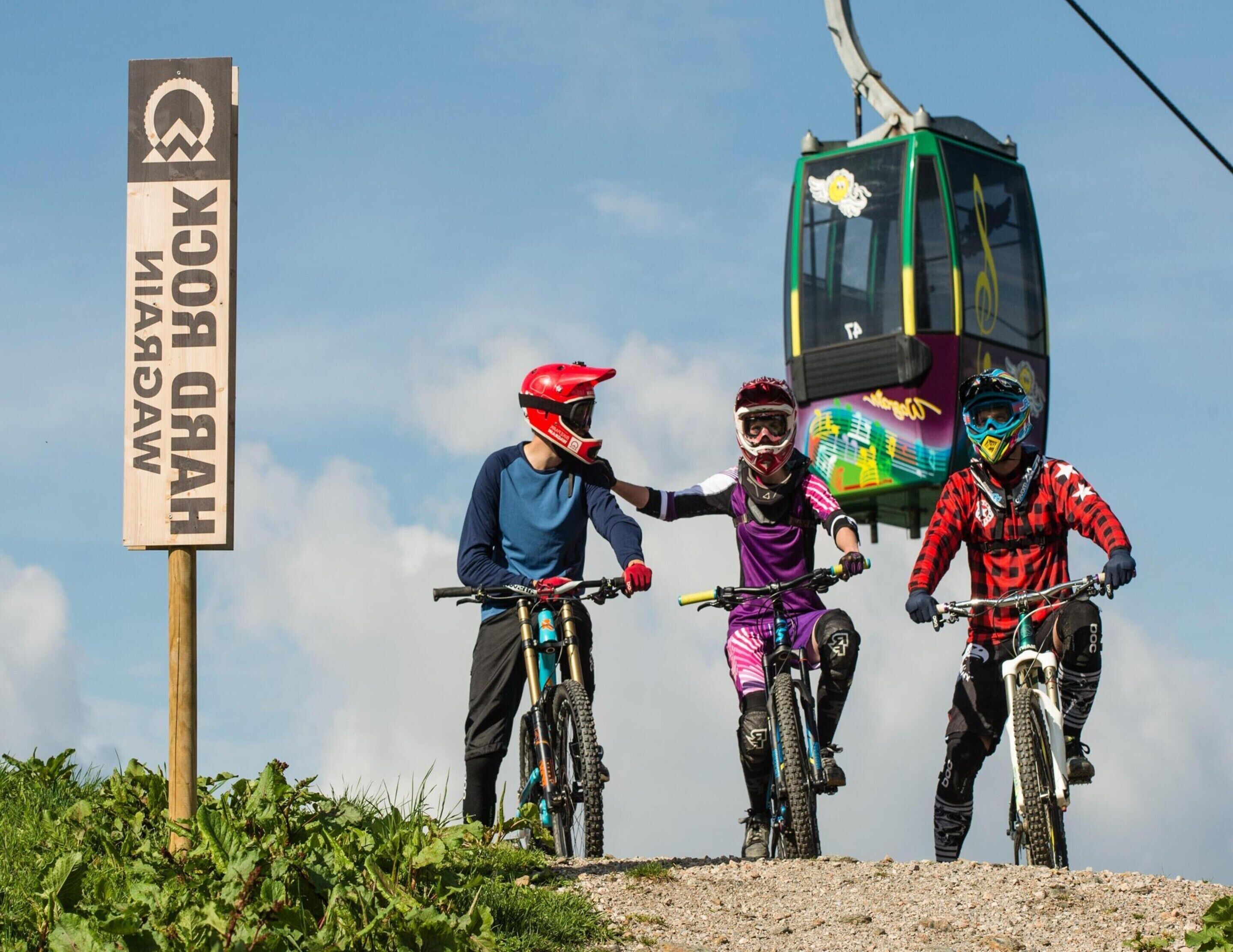 Three mountain bikers on a trail with a cable car in the background.