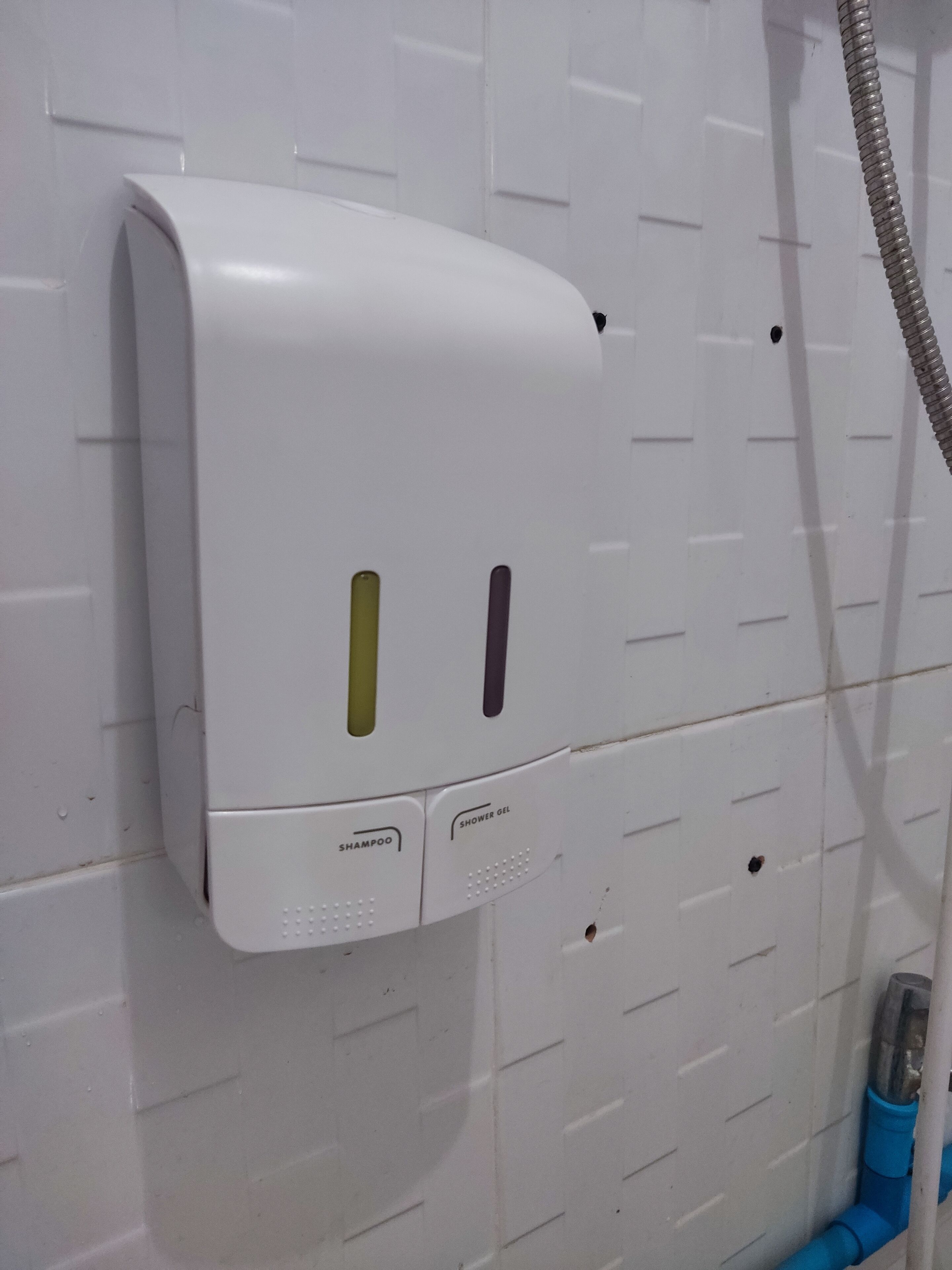 Ein weißer, an der Wand montierter Spender mit zwei Fächern, einem für Shampoo und einem für Duschgel, und einer blauen Leitung, die an der Wand verläuft.