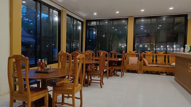 A dining area with wooden tables and chairs, large windows, and a seating area with a bench. - erawan guesthouse (Pakse)