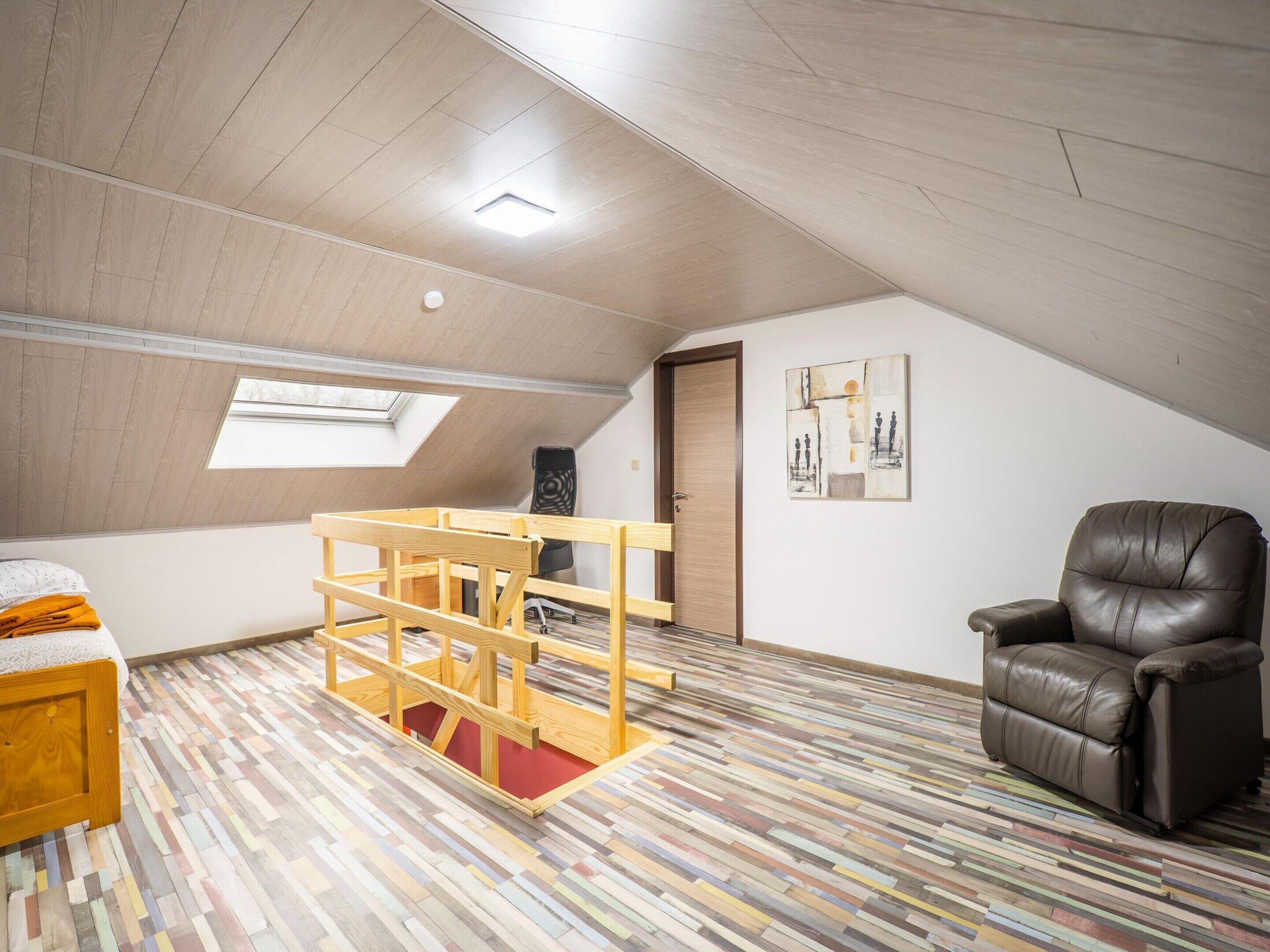 Un dormitorio tipo loft con techo inclinado, tragaluz, cama de madera, sillón de cuero marrón y piso con rayas de colores.