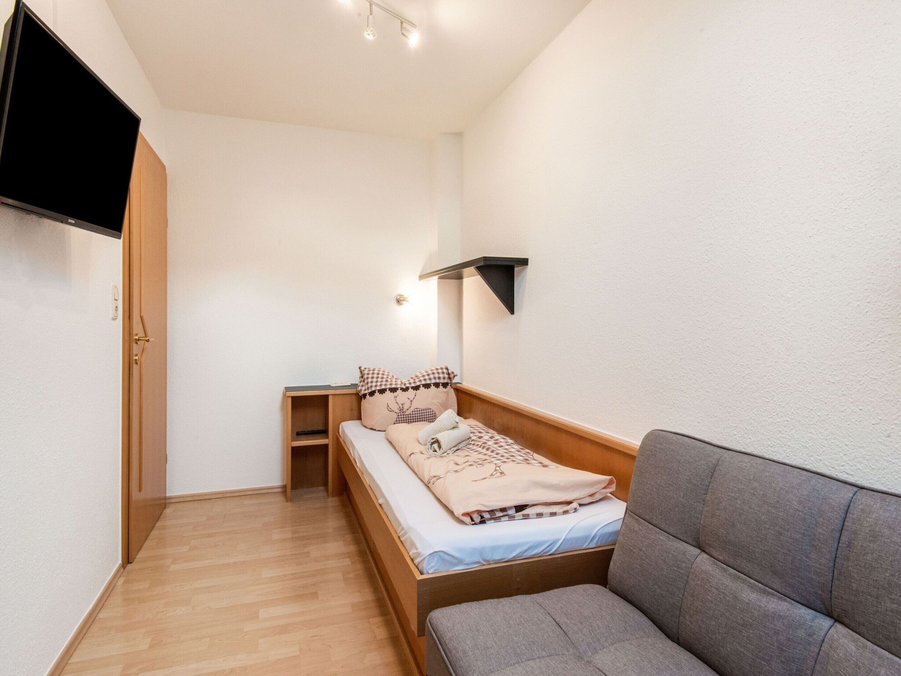 A small, neatly arranged bedroom with a bed, a sofa, a TV, and a shelf.
