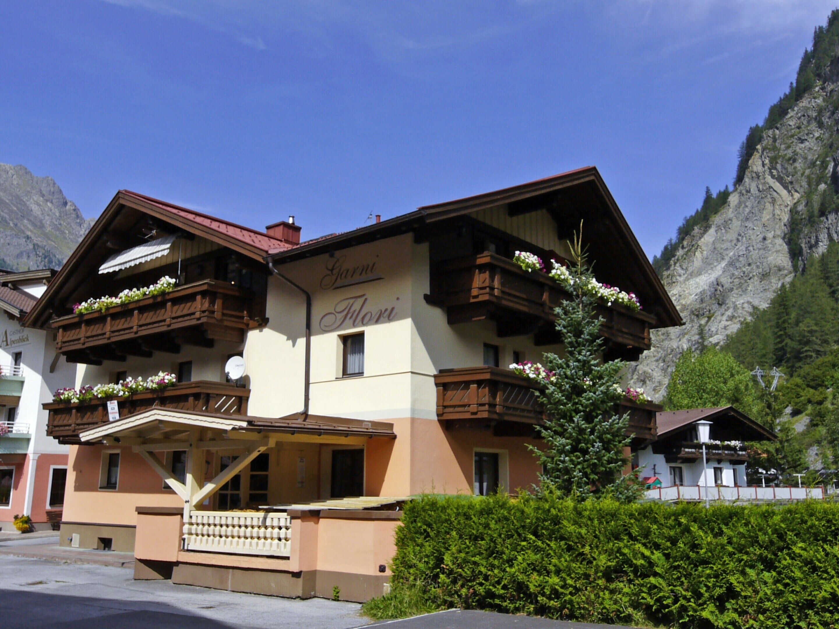 A hotel with balconies adorned with flowers, a sign reading "Garni Flori", and a mountainous backdrop.