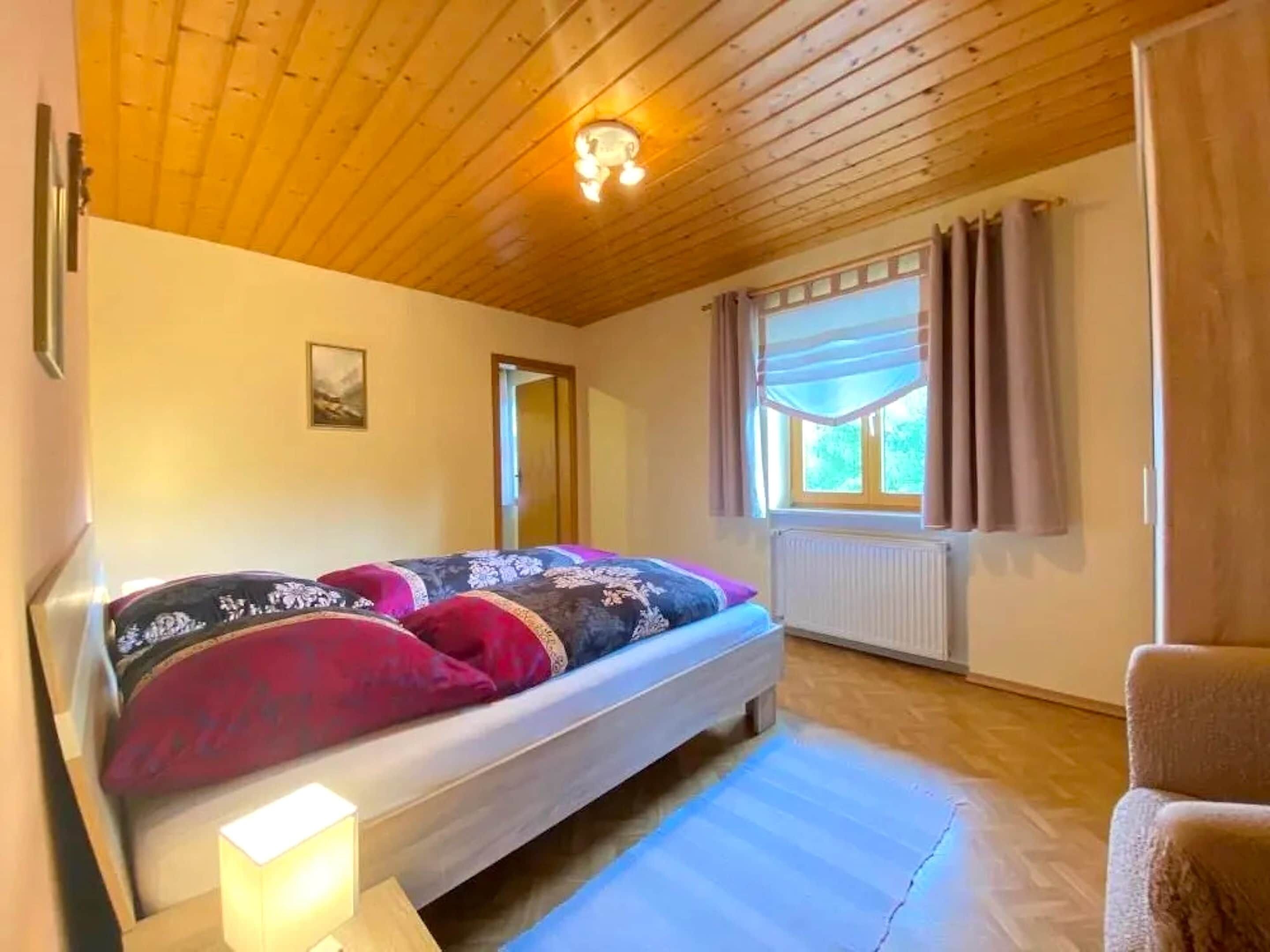 A bedroom with a wooden ceiling, a bed with patterned bedding, a window with curtains, and a radiator.