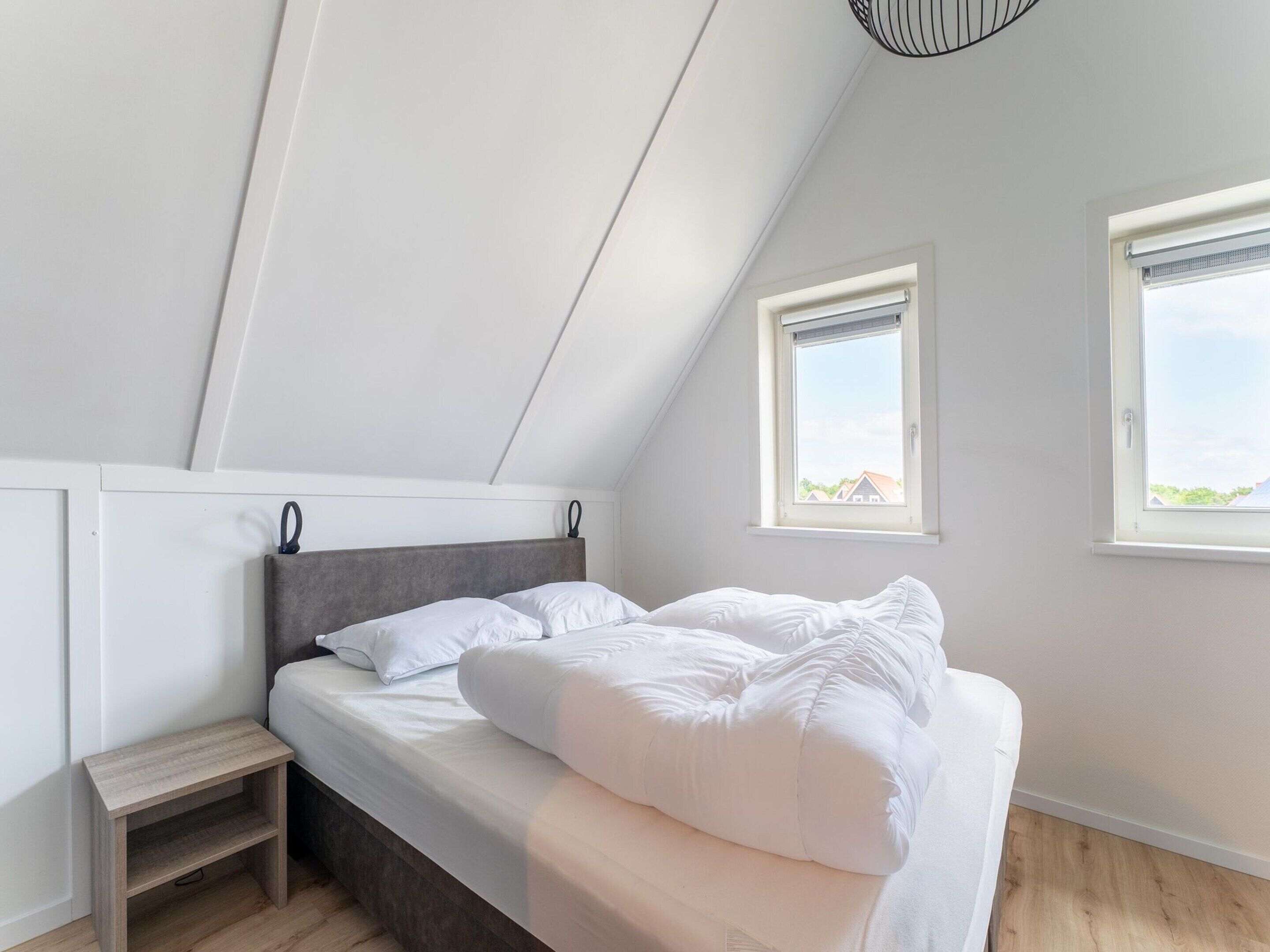 Une chambre avec un plafond en pente, un lit recouvert de linge de lit blanc, une petite table de chevet en bois et deux fenêtres donnant sur l’extérieur.