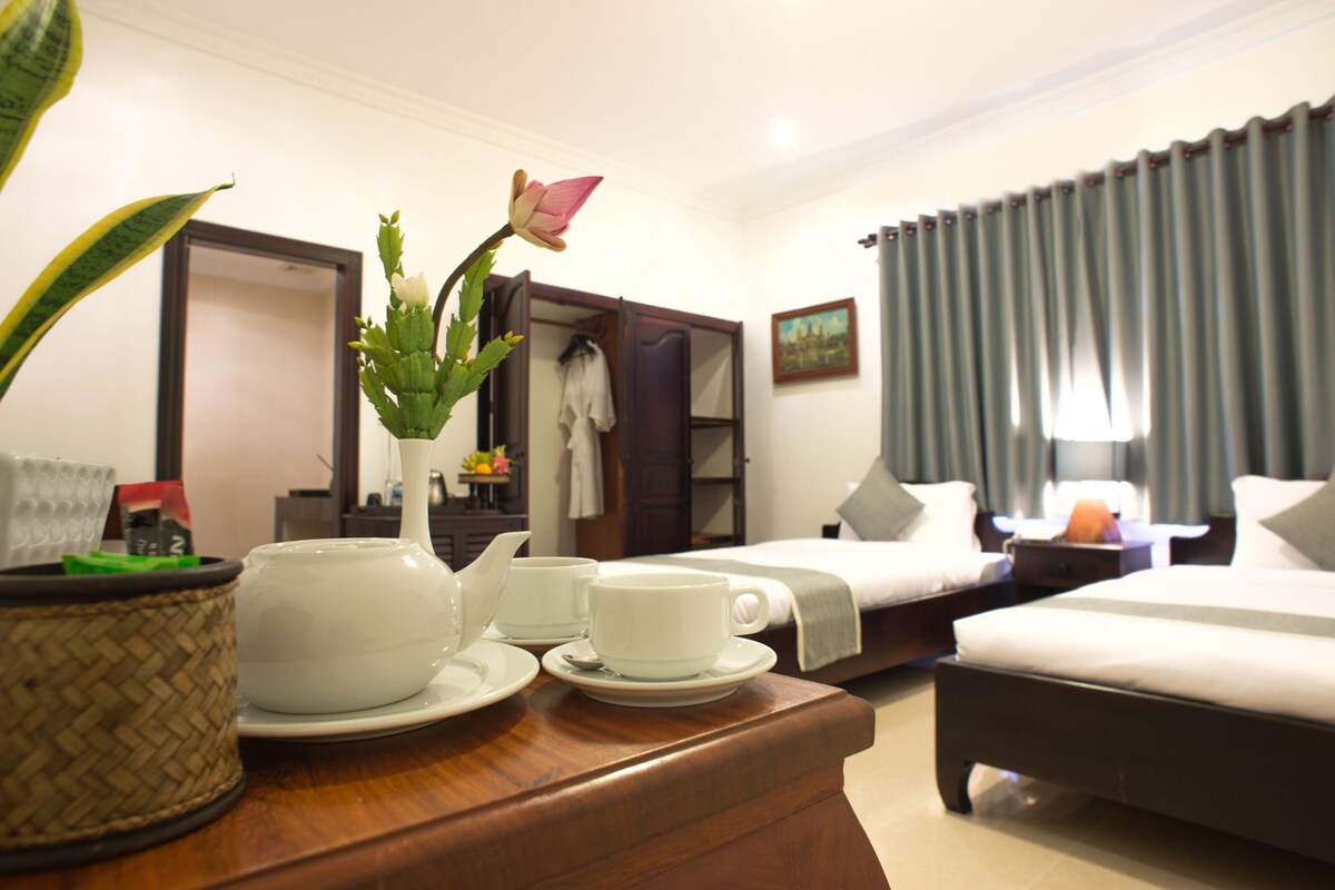A hotel room with a bed, a bedside table with a teapot, cups, and a potted plant.