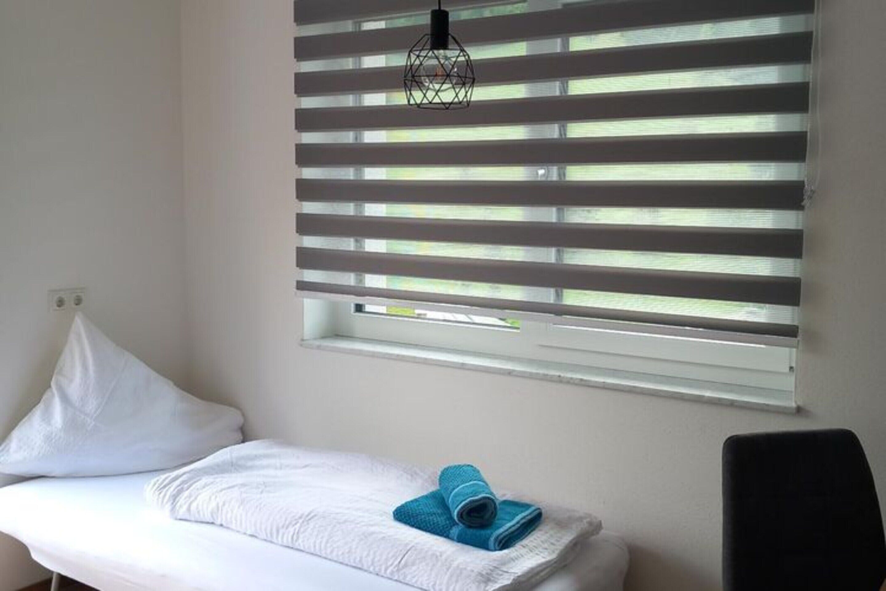 Una cama individual con ropa de cama blanca, una toalla azul y una silla negra en una habitación con paredes blancas y una ventana con persianas horizontales.