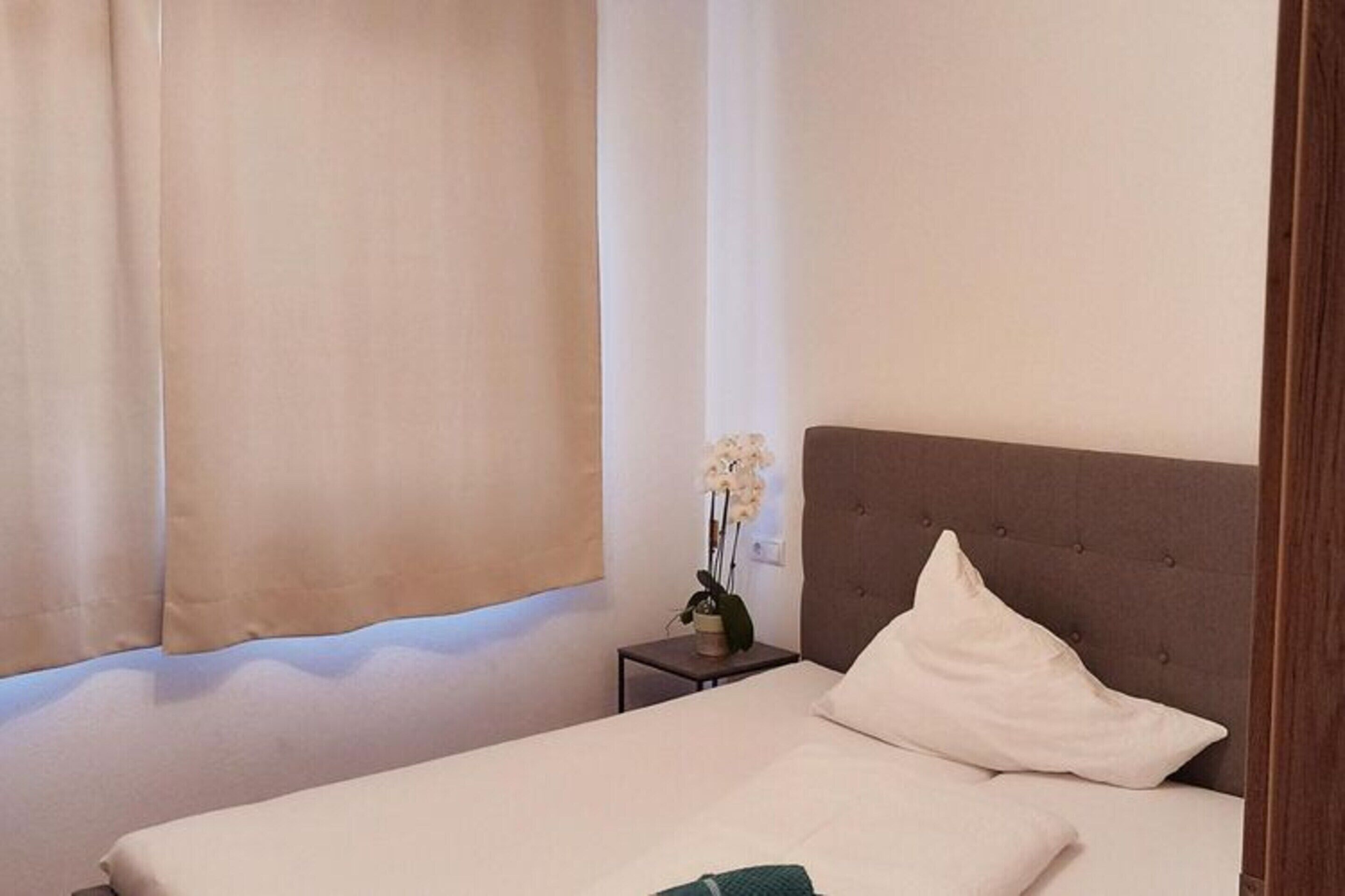 Una cama bien hecha con una almohada y una toalla verde, una mesita de noche con una planta, y una ventana con cortinas.