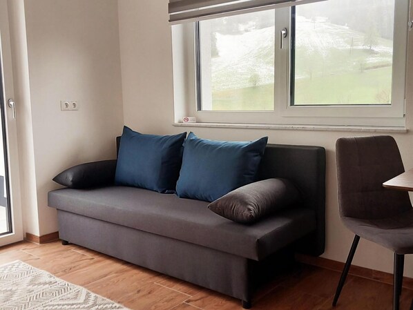 Sala de estar moderna com um sofá cinza, duas almofadas azuis, piso de madeira e uma janela com vista para uma paisagem verde.