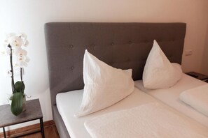 Cama bem arrumada com cabeceira estofada cinza, duas almofadas brancas e uma pequena mesa de cabeceira com uma orquídea em vaso.