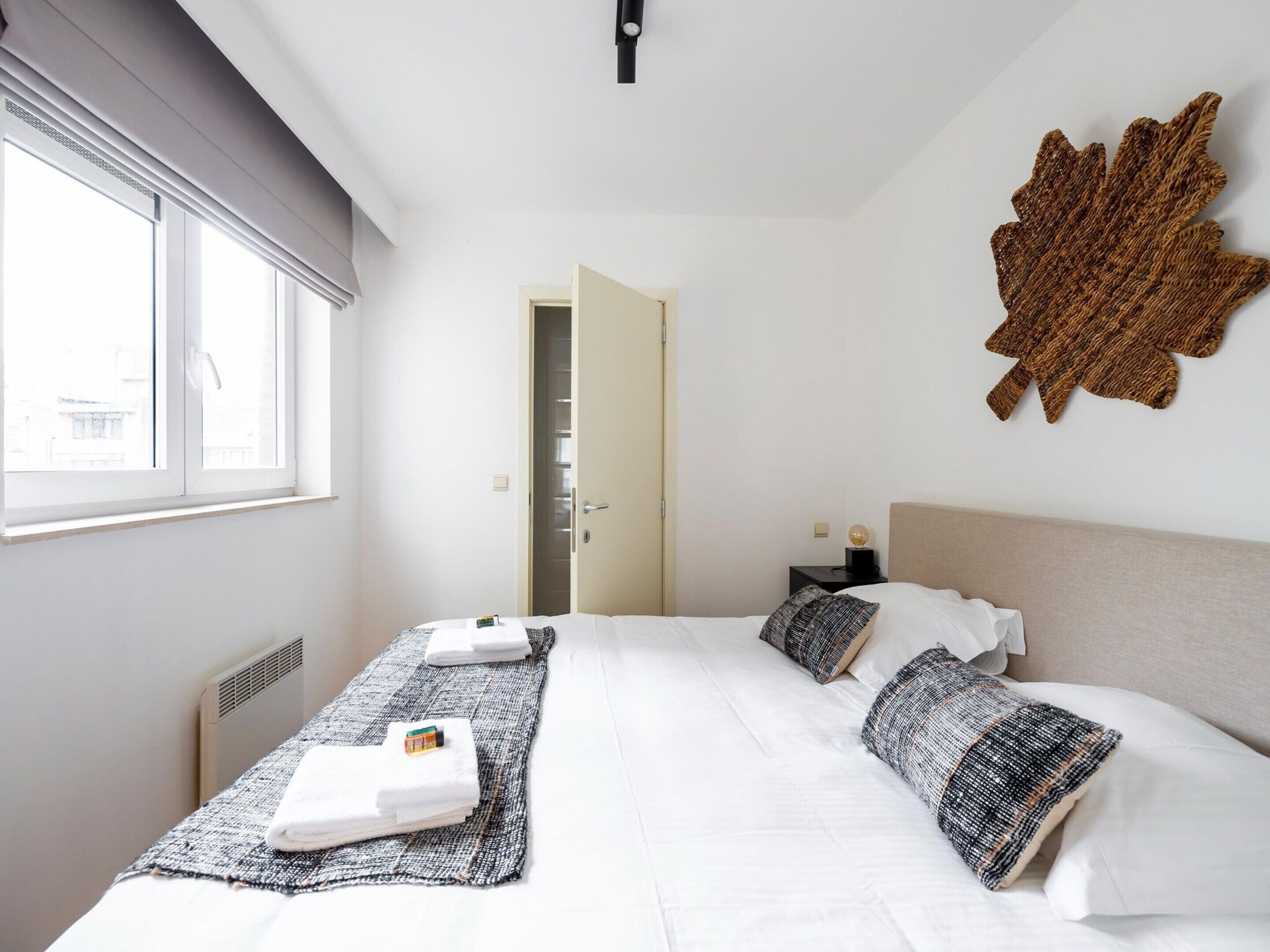 A hotel room with a bed, a bedside table, a wall-mounted decor piece, and a window with blinds.