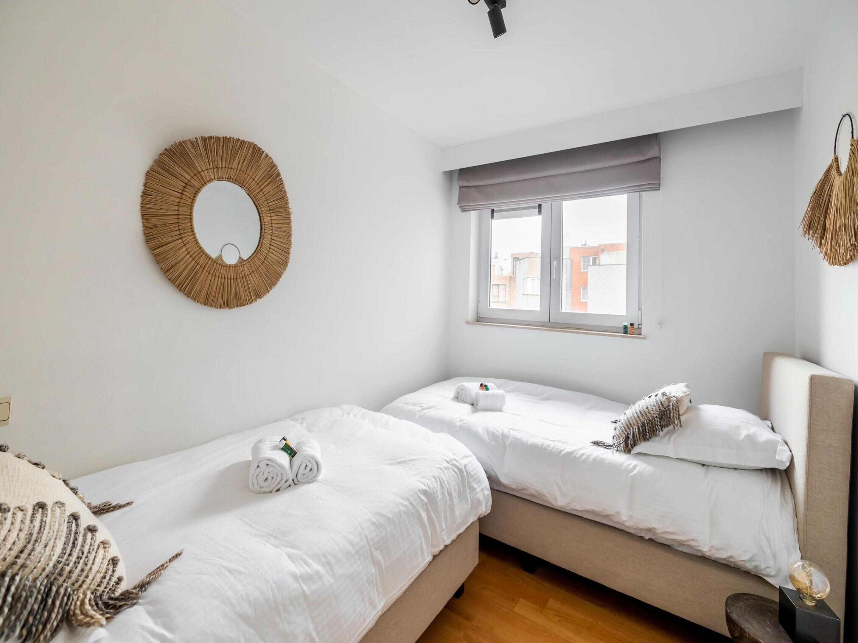 A hotel room with two single beds, a round mirror, a window with a view of buildings, and a hanging woven decoration.
